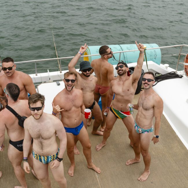 A group of men in colorful swimsuits are standing on a boat, smiling and posing for a photo. Some are holding drinks, and the sea is visible in the background.