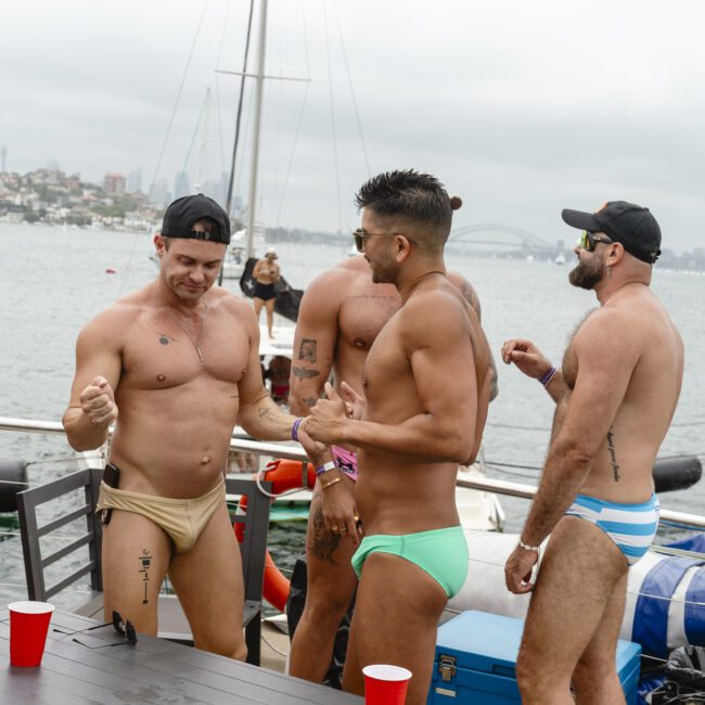 A group of men in swimwear socialize on a boat with a cityscape and bridge visible in the background. Red cups are on a table, and an inflatable safety buoy is attached to the boat.