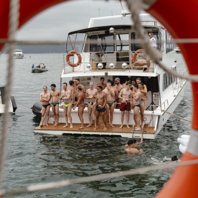 A group of people in swimsuits are standing on the deck of a boat, which is floating on the water. The scene is viewed through an orange life preserver. Another person is swimming in the water nearby. Several boats are visible in the background.