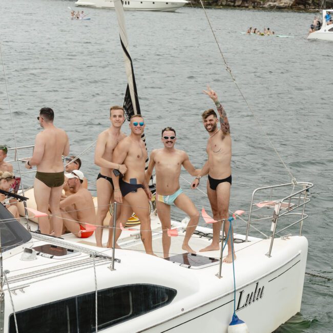 Four men in swimwear stand on a sailboat, smiling and waving. The boat is in a body of water, surrounded by other boats. Trees and land are visible in the background, suggesting a lively, social atmosphere.