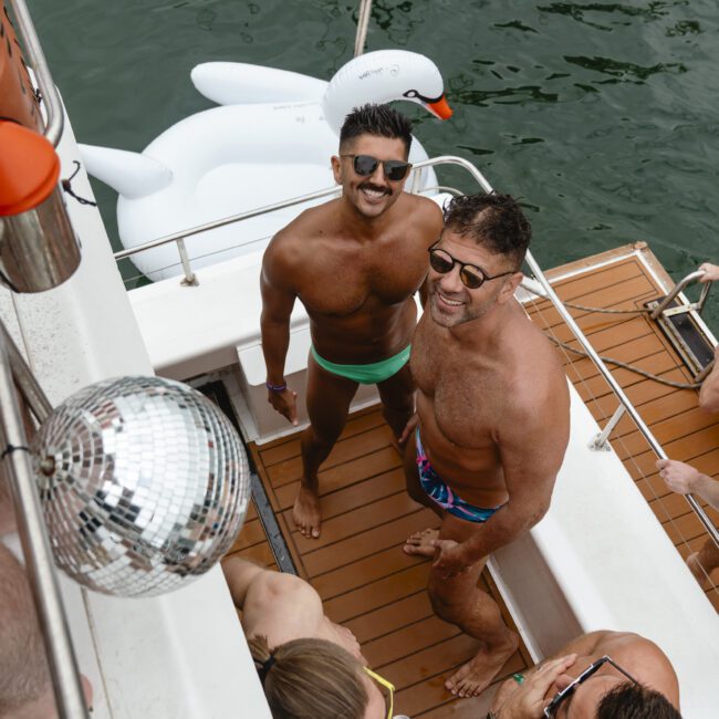 A group of people enjoying a boat party on a sunny day. Two men in colorful swimsuits are smiling at the camera. A white inflatable swan floats in the water nearby, and a disco ball hangs overhead.