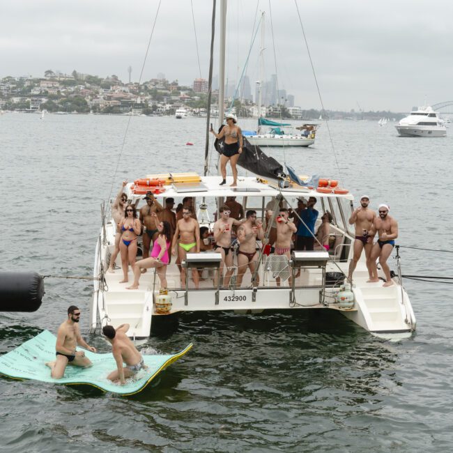 A group of people enjoying a day on a catamaran in a harbor. Some are standing on the deck, while others are on a large floating mat in the water. The background shows city buildings and more boats under a cloudy sky.