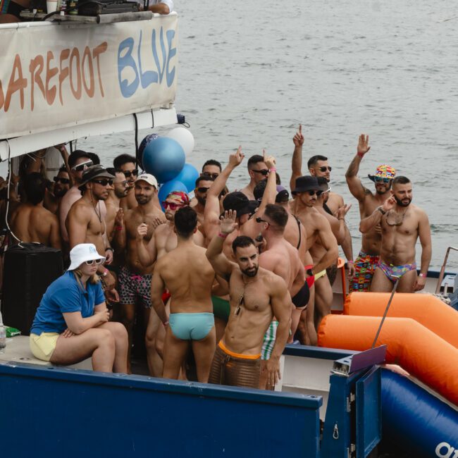 A group of men in colorful swimwear dance and socialize on a boat decorated with blue balloons. The boat is on a body of water with a cityscape in the background. Some men wave at the camera, while others chat and enjoy the event.