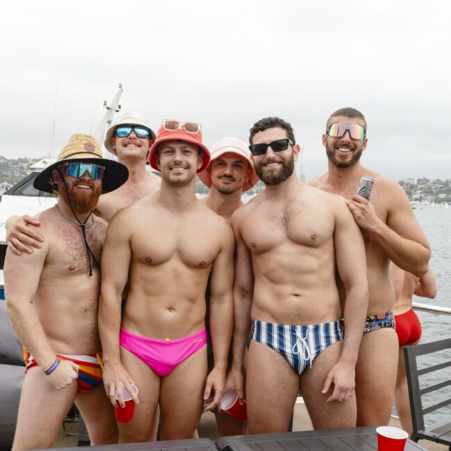 A group of six men in colorful swimwear pose together on a boat. They are smiling, and some wear sunglasses and hats. In the background, a body of water and another boat are visible.