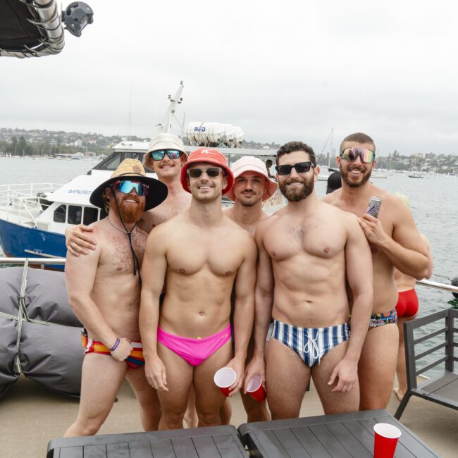 Six men in swimwear pose on a boat deck. They wear hats and sunglasses, holding red cups. The background shows a cloudy sky and water with boats.