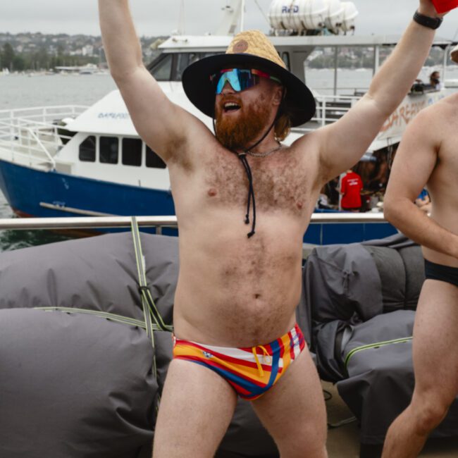 A man wearing a colorful swimsuit, sunglasses, and a wide-brimmed hat energetically poses with arms raised, holding a red cup. Behind him is a boat on the water, and cloudy skies cast a muted light over the scene.