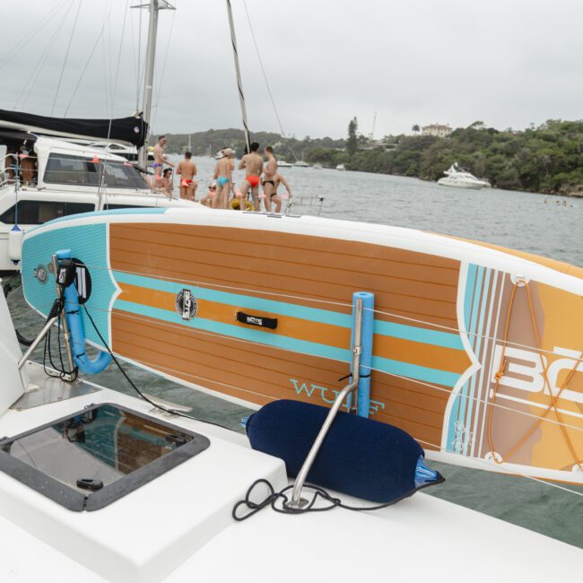 A stand-up paddleboard secured on a boat's side, with a group of people sitting on another boat in the background. The scene is set on a cloudy day in a calm, lush bay with several anchored boats.