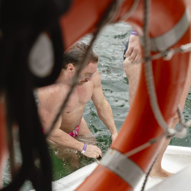 A person wearing swim trunks climbs on a boat, framed by a life preserver in the foreground. The water is visible behind them, indicating they have just emerged from swimming.