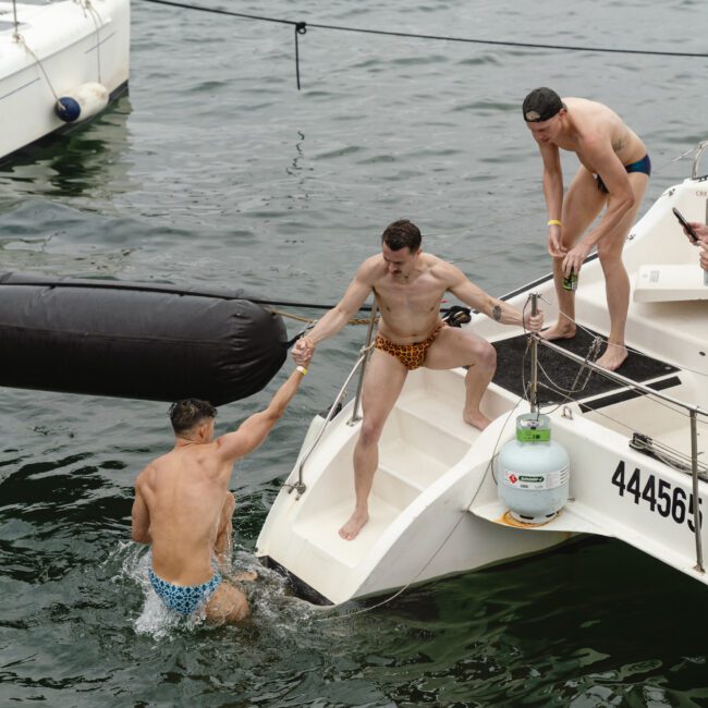 Three men on a boat, one in the water, wearing swim trunks. One man in leopard print trunks helps the swimmer back onboard. Another man stands nearby with a drink. Water and a docked sailboat are visible in the background.