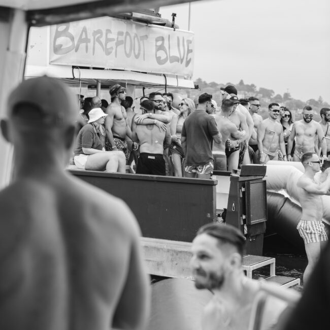 A group of people in swimwear gather on a deck labeled "Barefoot Blue" on a sunny day. Some are seated while others stand, with a man watching the scene in the foreground. Inflatable boats are visible in the background.