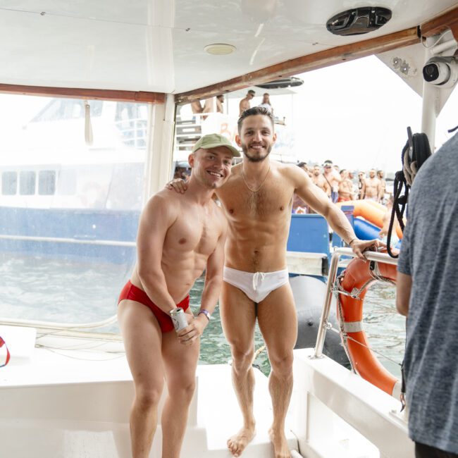 Two men in swimwear stand on a boat, smiling at the camera. One wears a red swimsuit and cap, the other a white swimsuit. A crowd is visible on another boat in the background, with water all around.