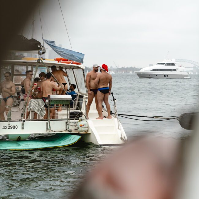 A group of people on a boat, some wearing swimsuits, with one person in a Santa hat. They are anchored near a yacht in a cloudy sky, with a paddleboard visible on the water.