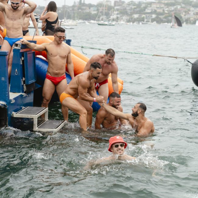 A group of men in swimwear are helping each other get out of the water onto a pontoon, with an inflatable corner visible. One man in the foreground wears a red bucket hat and sunglasses, smiling in the water. Boats and a shoreline are seen in the background.