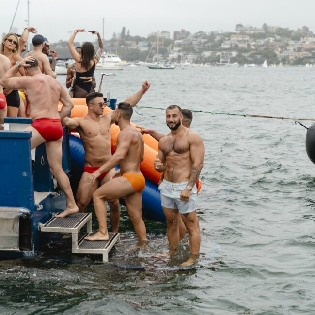 A group of people in swimwear enjoy a boat party. Some are standing in the water near the boat, while others are on the boat deck. The background shows a cloudy sky and a town across the water.