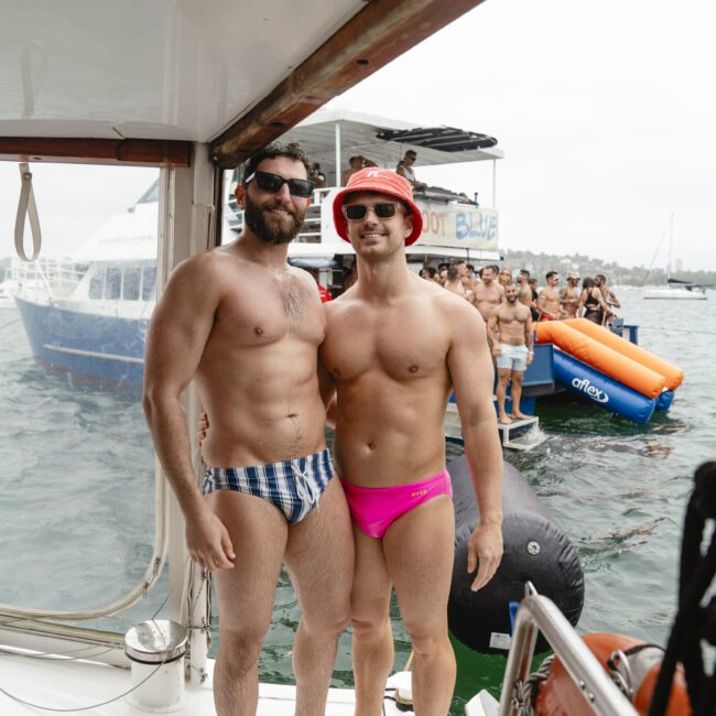 Two men in swimwear stand on a boat, smiling at the camera. The man on the left wears checkered swim trunks, while the man on the right wears pink swim trunks and a red bucket hat. Other boats and people are visible in the background on the water.