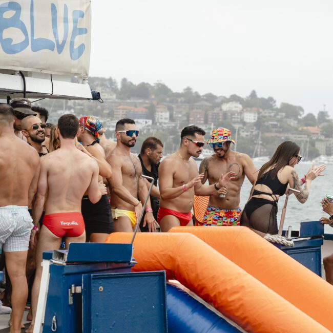 A group of people in swimwear socialize and enjoy drinks on a boat with a slide. The scene is lively, featuring colorful swim trunks and a cityscape in the background on a cloudy day.