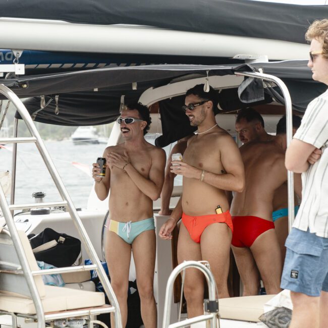 A group of four men are standing on a boat, wearing colorful swimwear. Two are holding canned drinks and appear to be laughing. Another man is standing nearby in a striped shirt. The boat is anchored with a view of water in the background.