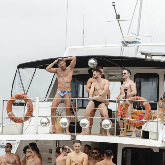 A group of men wearing various swimwear gather on the deck of a boat, some posing and interacting. Life buoys are visible on the railing. The scene suggests a lively atmosphere under a cloudy sky.