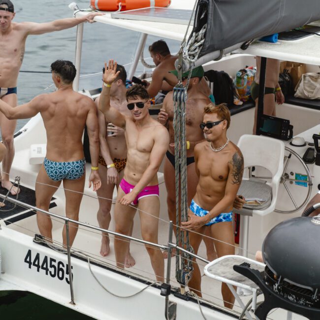 A group of men in swimsuits stand and relax on a boat deck in the water. Some are waving and wearing sunglasses, while another leans on the railing. The boat is equipped with safety gear and a partial canopy. The background shows the water.