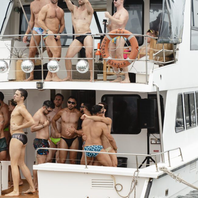 A group of men in swimwear are gathered on the deck of a yacht. Some are standing and posing while others chat and relax. The scene suggests a festive, sunny day spent on the water.