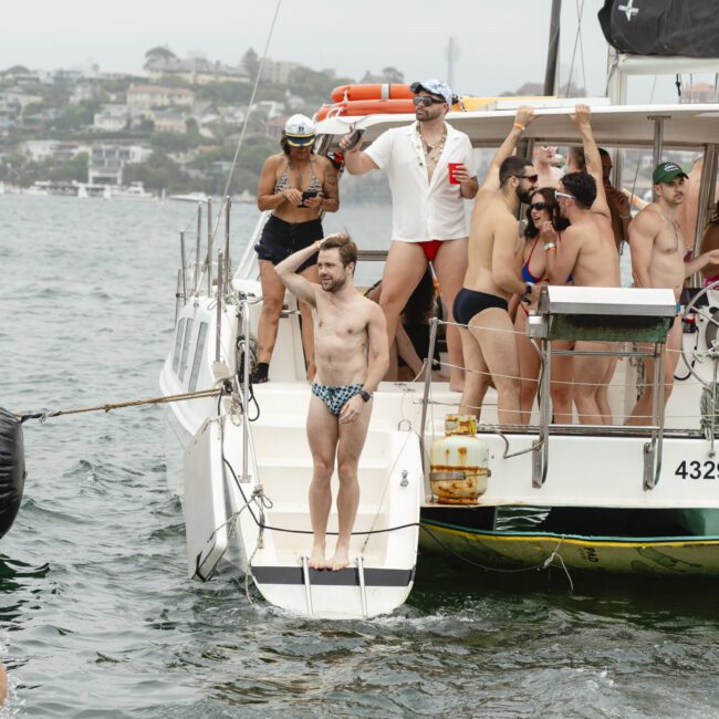 A group of men in swimwear stand on a yacht, some holding drinks and a speaker. One man is at the edge ready to dive into the water. The yacht is on a calm, overcast day with a shoreline and houses visible in the background.