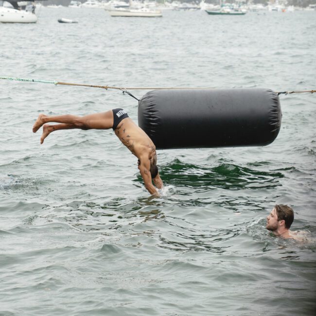 A man in swim trunks is mid-dive from a boat into the water, with another man swimming nearby. Several boats are visible in the background on a cloudy day.