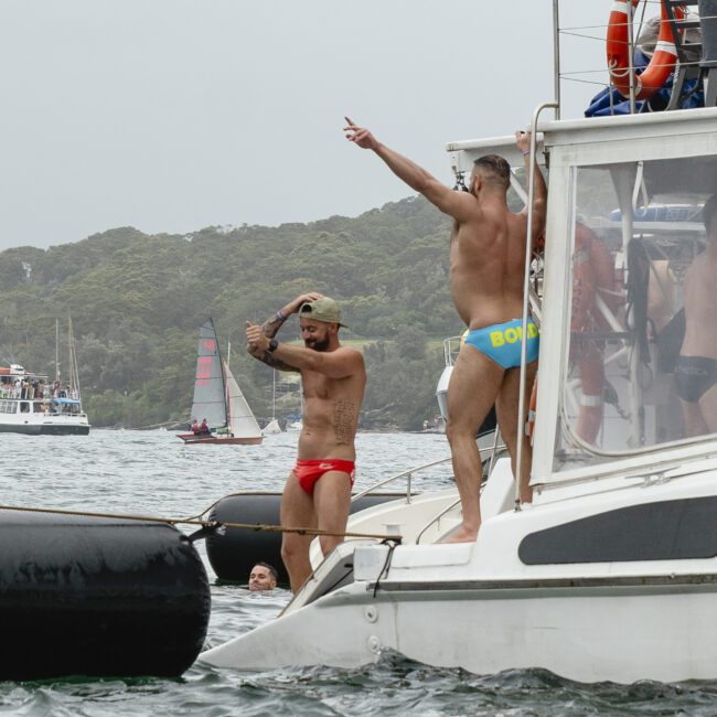 Two men in colorful swimwear stand on the edge of a boat, preparing to jump into the water. The boat is surrounded by other vessels in a scenic bay with forested hills in the background. One wears red, the other blue with "Bondi" written on it.