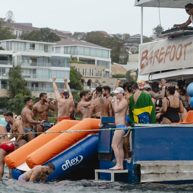 A lively boat party with people dancing and enjoying themselves. Some are wearing swimsuits, others are on inflatable rafts. A DJ plays music under a banner that reads "BAREFOOT BLUE." A person waves a Brazilian flag.