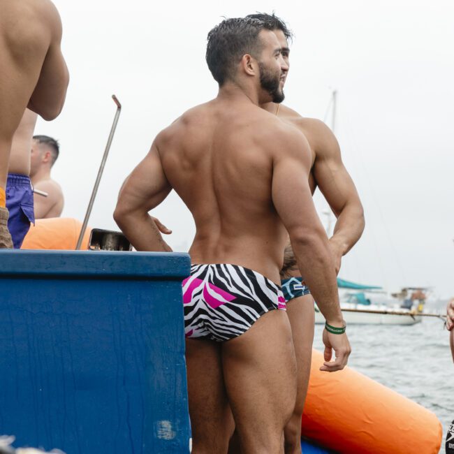 Two men in swim trunks stand on a dock by the water. One wears a colorful zebra-patterned trunk. The scene shows a cloudy day with a boat in the background.