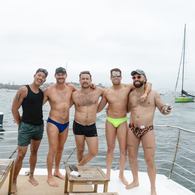 Five men wearing swimwear stand on a boat's deck, posing for a photo against a backdrop of a cloudy sky and sailboats on the water. They appear relaxed and smiling, enjoying their time by the sea.