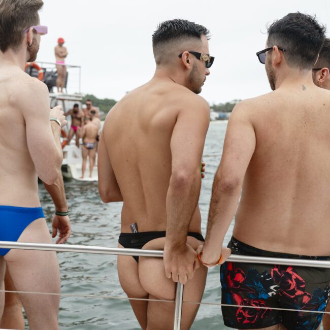 A group of men on a boat, some shirtless and wearing swimwear. They are standing near a railing, with water in the background. It appears to be a casual, social setting on a cloudy day.