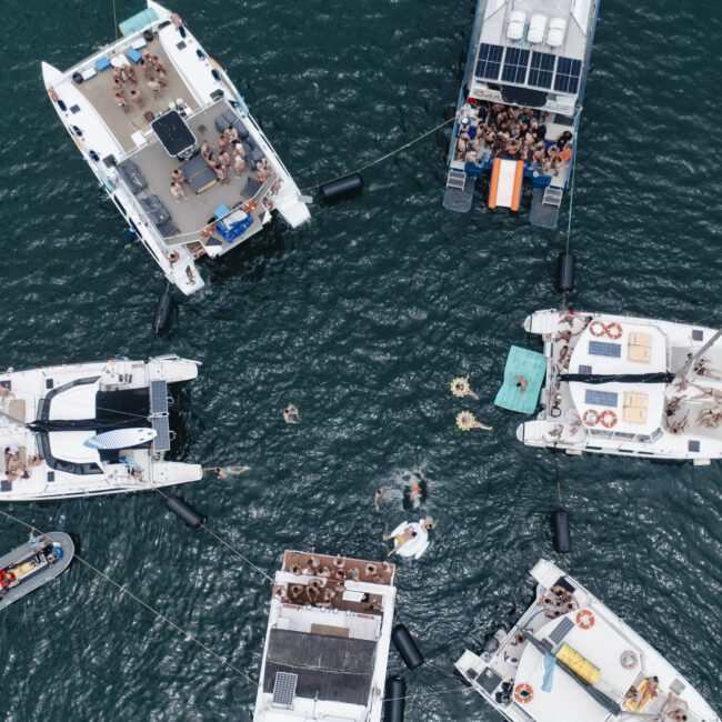 Aerial view of six sailboats and a dinghy gathering in a circle on the water. People are sunbathing, socializing, and swimming. Inflatable floats are visible between the boats. The scene captures a lively boating event.