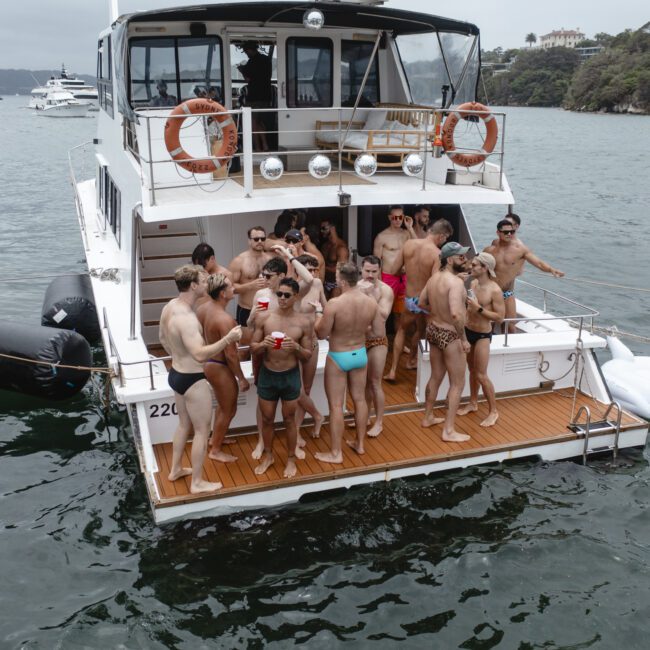 A group of people on the back deck of a boat are enjoying a party. They are wearing swimsuits and holding drinks. The boat is on a calm body of water, and there's a large inflatable swan floating nearby. Other boats are visible in the background.