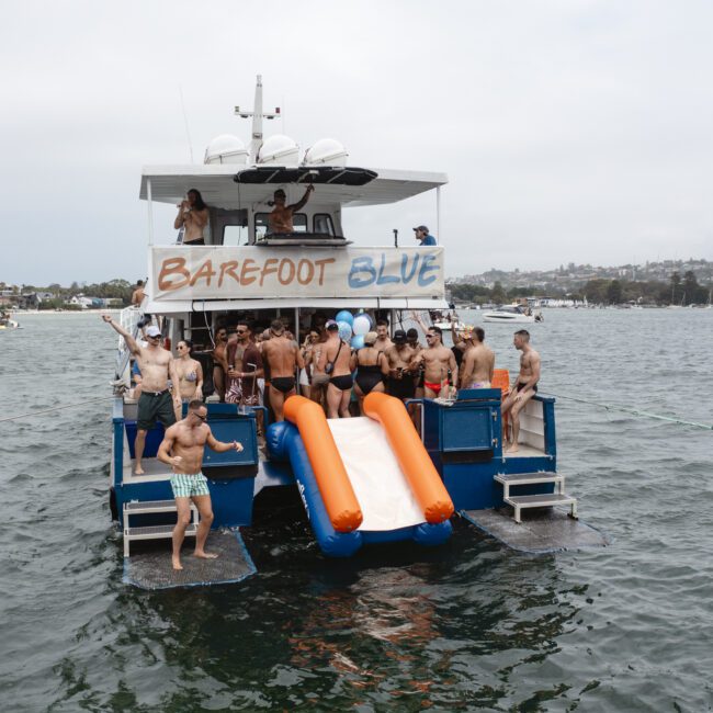 A group of people in swimwear are gathered on a boat named "Barefoot Blue" with inflatable slides at the back. The boat is on a calm body of water with a shoreline and other boats visible in the background.