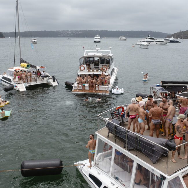 A group of people enjoying a party on multiple boats anchored in a bay. Several people are wearing swimwear and socializing, with overcast skies in the background. Other boats are visible in the distance.