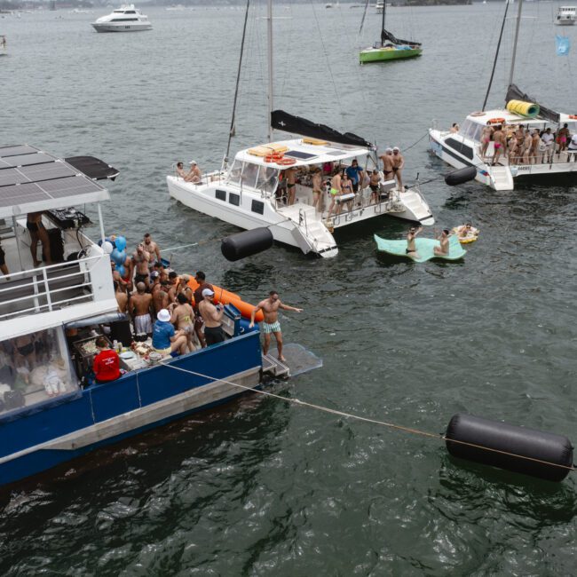 Boats filled with people are anchored close together on a body of water, with individuals swimming and socializing. Floating platforms and inflatable barriers are visible between the boats. It's a lively scene suggesting a party or gathering.