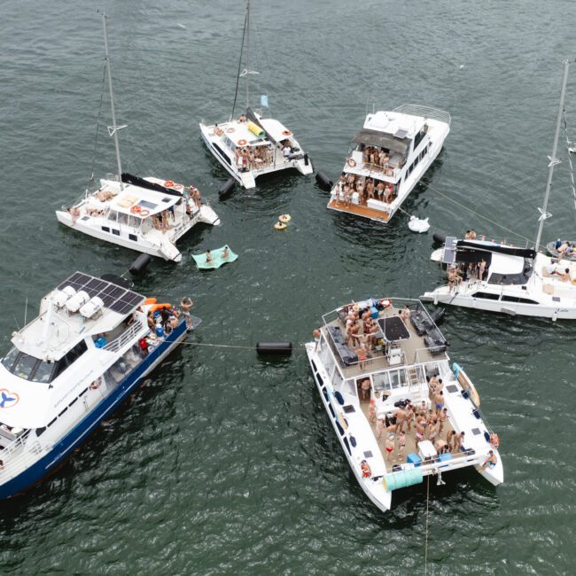 Aerial view of several boats gathered in a circle on the water. People are socializing on the decks, some on floaties between the boats. The water is calm, and the sky is clear, suggesting a sunny day.