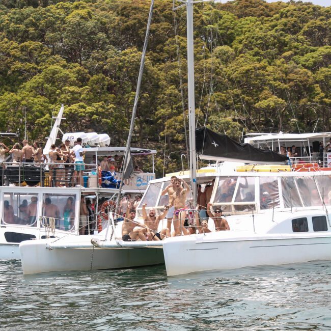 A group of people are gathered on several boats anchored near a wooded shoreline. They are enjoying a sunny day on the water, with some standing and others sitting. The scene suggests a lively atmosphere of leisure and socializing.