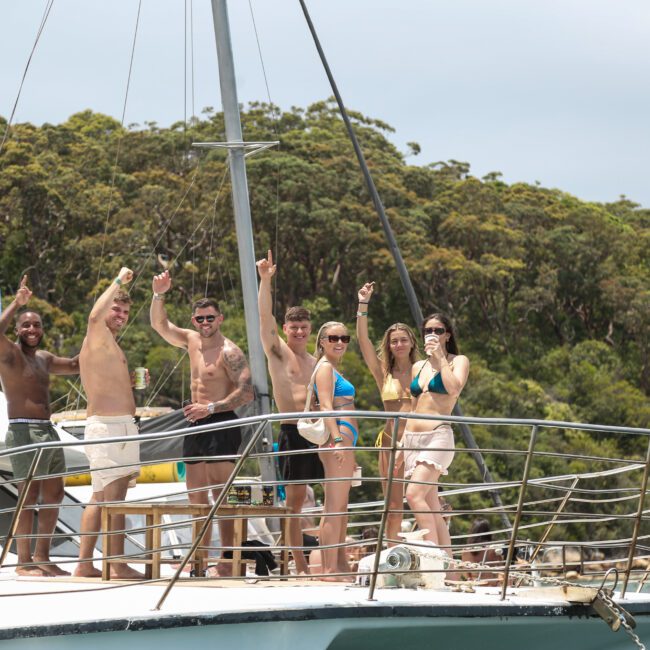 A group of people in swimwear happily posing on the deck of a boat. They are surrounded by lush green trees and water, enjoying a sunny day. Some are raising their arms in celebration.