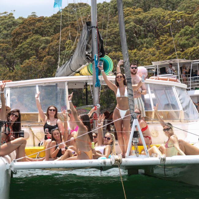 A group of people in swimsuits enjoying a sunny day on a sailboat, surrounded by lush green trees. They are laughing, posing, and raising their arms in celebration, with drinks in hand. Several boats are nearby in the background.