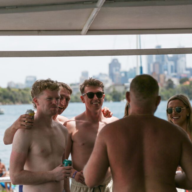 A group of people on a boat, with some shirtless men and a woman wearing sunglasses, smiling and enjoying drinks. The background shows a city skyline and water, suggesting a sunny day.