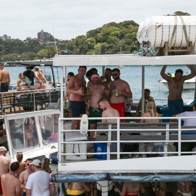 A group of people enjoying a sunny day on a party boat. Some are standing on the upper deck, holding drinks and smiling, surrounded by water and trees in the background. Other passengers are visible on the lower deck and nearby boats.