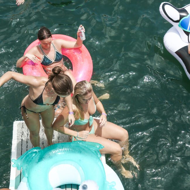 Four people in swimsuits enjoy lounging on inflatable pool floats in clear water. One woman balances on a float while another laughs nearby. The scene suggests a fun, sunny day at the pool or lake.