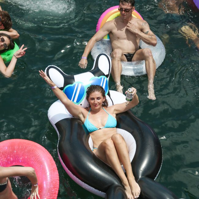 A woman in a blue bikini lounges on a black inflatable cat float, holding a can, while a man in pink sunglasses relaxes nearby on a donut-shaped float. Others are enjoying the water around them on a sunny day.