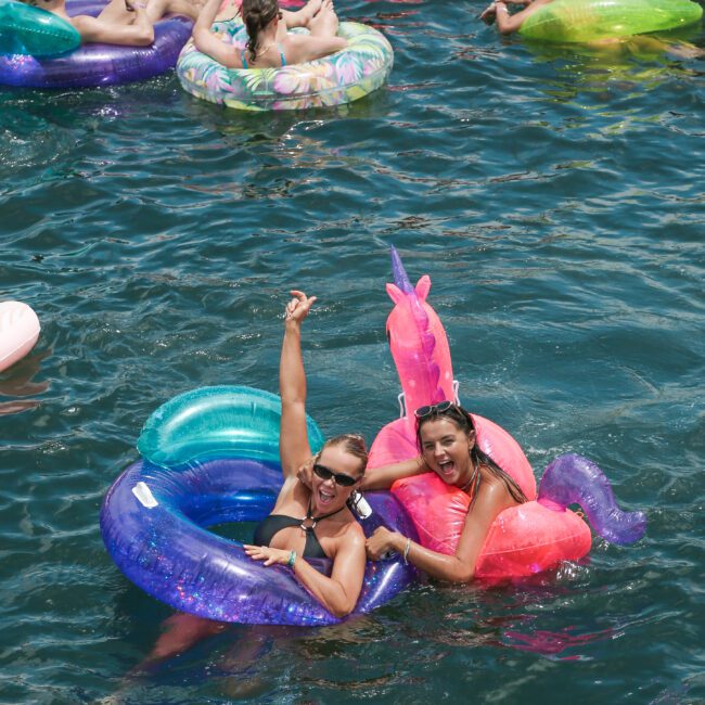 Two people in colorful inflatable floats, one a unicorn, smiling and waving while enjoying a sunny day in the water. Others on floats are visible in the background.