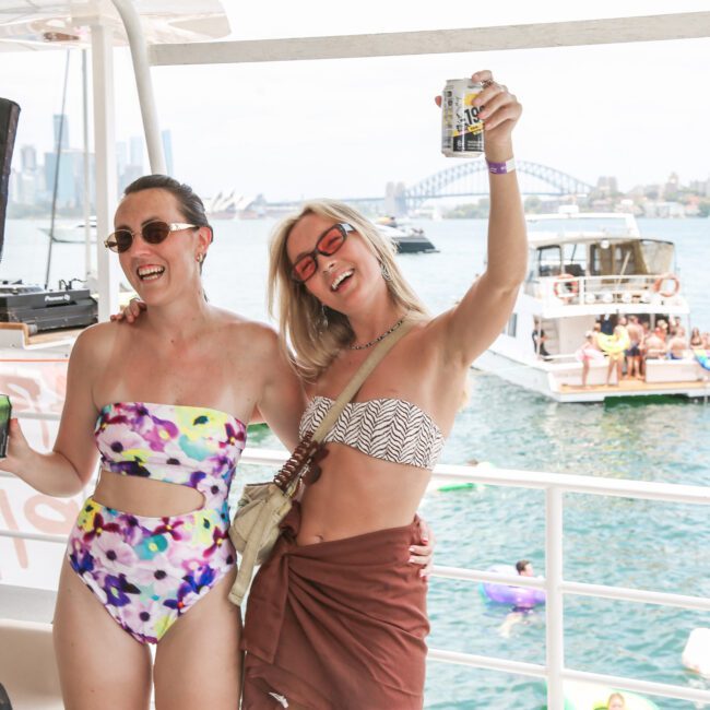 Two people in colorful swimsuits enjoy a boat party, holding drinks and smiling. The background shows a view of the water with other boats and people swimming, and a city skyline in the distance.