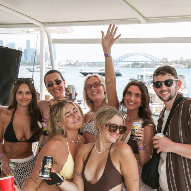 A group of eight people enjoying a sunny day on a boat, holding drinks and smiling. The background features a body of water, city skyline, and a bridge. Everyone is wearing casual summer attire, including swimsuits and sunglasses.