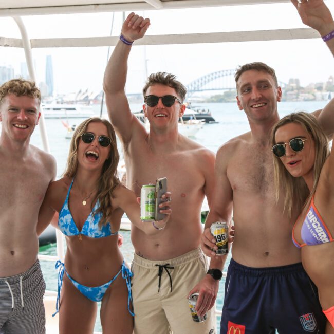 A group of five people in swimwear poses happily on a boat. They're holding drinks and smiling, with a city skyline and a bridge visible in the background.