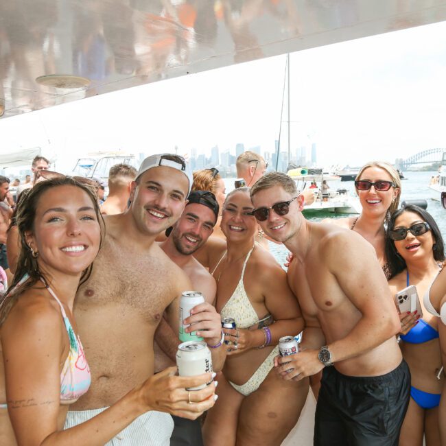 A group of people in swimsuits are smiling and holding drinks on a boat. It's a sunny day, and other boats and city skyline can be seen in the background.