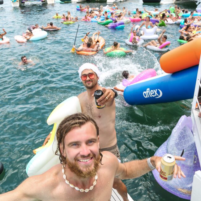 A lively scene with people enjoying a boat party in the water. Two men in swimwear hold drinks, smiling at the camera. Numerous inflatables and colorful pool toys float around, with a crowd and boats in the background.